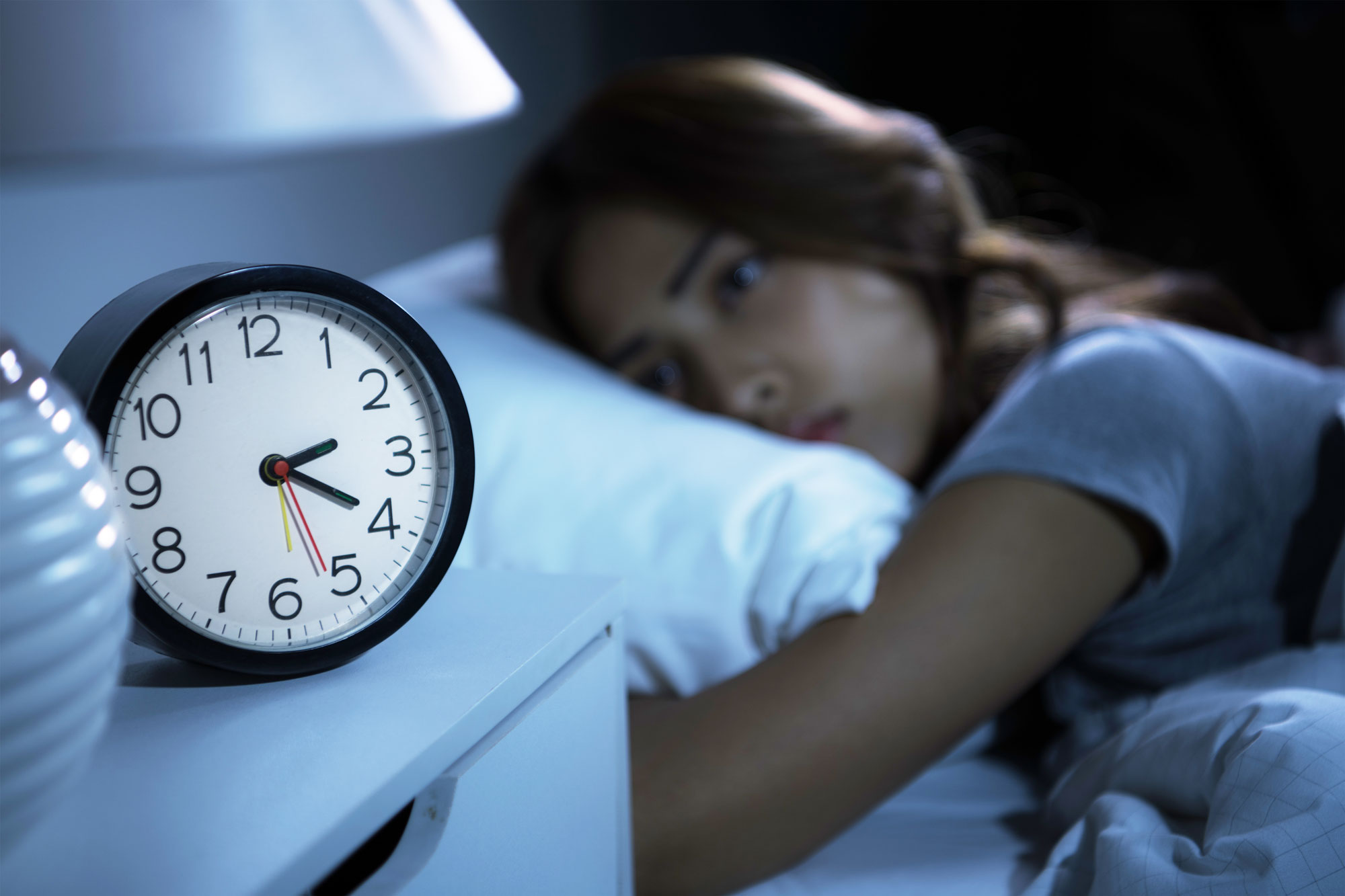 Frau liegt schlaflos im Bett und schaut auf Wecker der neben dem Bett steht.